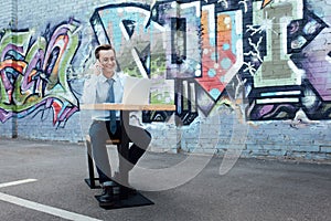 smiling young freelancer in eyeglasses talking by smartphone and using laptop while sitting