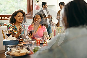 Smiling young friends drink wine and looking on phone during holiday party at home