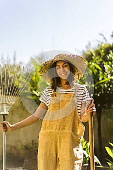 Smiling young gardener with straw hat with rake and shovel in her hands. Gardening.