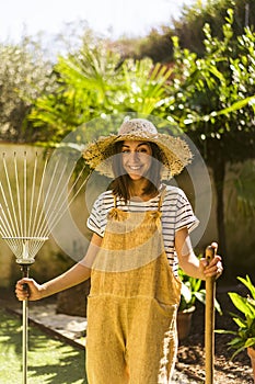 Smiling young gardener with straw hat with rake and shovel in her hands. Gardening.