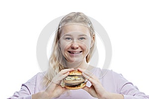 Smiling young girl with a big hamburger in her hands. Cute blonde teenager with freckles in a pink sweater. Delicious garbage food