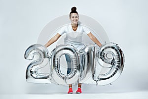 Smiling young girl dressed in white t-shirt, jeans and pink socks holding balloons in the shape of numbers 2019 on the