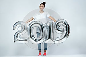 Smiling young girl dressed in white t-shirt, jeans and pink socks holding balloons in the shape of numbers 2019 on the