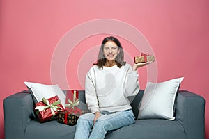 Smiling young girl hold festive gift box, showing present on palm, sitting on couch with holiday gifts boxes