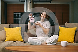 Smiling young girl hold mechanical spring alarm clock sitting on comfortable couch, notify about deadline or time frame