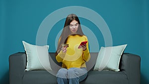 Smiling young girl holding bank credit card, phone sitting on couch, making purchase in online store. E-commerce