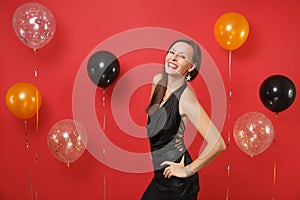 Smiling young girl in little black dress celebrating standing with arms akimbo on bright red background air balloons. St