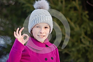 Smiling young girl in pink coat shows ok , against green of winter park