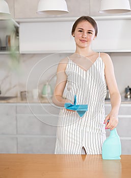 Smiling young girl standing at the table holding dustcloth and spray detergent