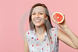 Smiling young girl in summer clothes holding in hand half of fresh ripe grapefruit isolated on pink pastel wall