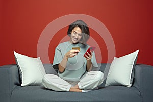 Smiling young girl use phone app to make easy payments online, hold credit card sitting on sofa. E-commerce, web banking