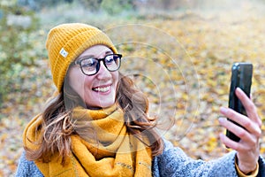 Smiling young girl in a yellow hat and scarf takes selfie in the autumn park. Close-up. Selective focus