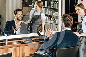 Smiling young hairdresser looking at handsome happy client showing ok sign and looking at mirror