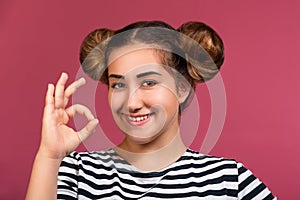 Smiling young hipster girl with playful hairstyle making ok hand gesture isolated over pink background