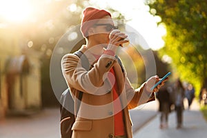 Smiling hipster man wear sunglasses using cellphone standing at the street drinking takeaway coffee