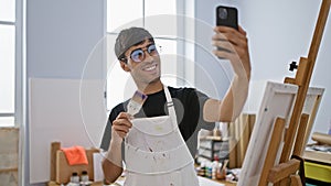 Smiling young hispanic man artist taking a splendid selfie picture with his smartphone at the bustling art studio, canvas in one