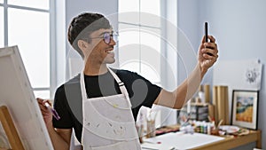 Smiling young hispanic man artist taking a splendid selfie picture with his smartphone at the bustling art studio, canvas in one