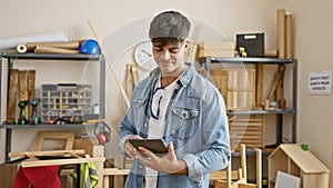 Smiling young hispanic man, a professional carpenter, engrossed in his carpentry work indoors, skillfully using a touchpad in his
