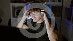 Smiling young hispanic man streamer confidently playing video game, wearing headphones, in his gaming room, immersed in digital