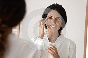 Smiling young hispanic woman apply nourishing cream under eyes