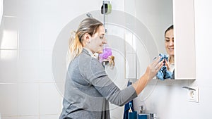 Smiling young housewife polishing mirror in bathroom above sink