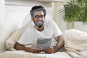 Smiling Young Indian Man With Digital Tablet Resting In Bed At Home