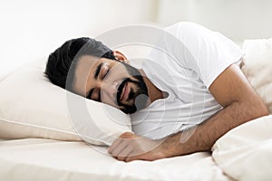 Smiling Young Indian Man Sleeping Calmly In Comfortable Bed At Home
