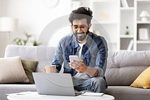 Smiling Young Indian Man Using Smartphone And Drinking Coffee At Home