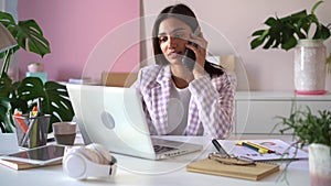 Smiling young Indian mixed-race business woman professional talking on phone using laptop sit at office desk