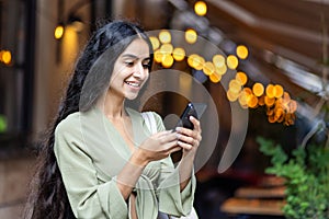 A smiling young Indian woman in a green suit stands on an evening city street and uses a mobile phone in her hands and