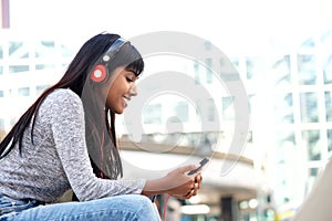 Smiling young Indian woman sitting outside in the city listening to music with mobile phone and headphones