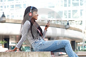 Smiling young Indian woman sitting outside in the city listening to music with smart phone and headphones