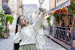 Smiling young Indian woman traveling and walking on city street, standing and taking selfie on mobile phone, showing