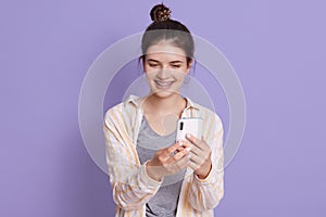 Smiling young lady with hair bun holding modern smart phone in hands and making selfie, looking smiling at device screen, standing