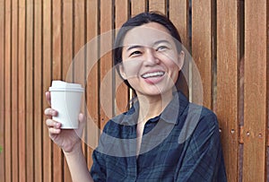Smiling young lady standing at the wooden wall outdoors and holding a cup of coffee