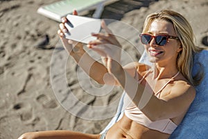 Smiling young lady on vacation taking photos using smartphone while relaxing on the beach