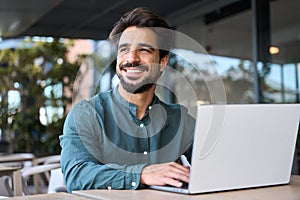 Smiling young Latin business man using laptop working outdoors looking away.