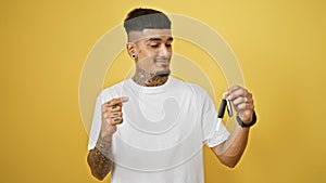 Smiling young latin man confidently pointing to key of his new car, isolated on yellow background