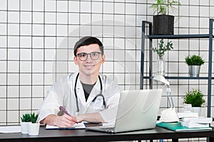 Smiling young male doctor working at the clinic reception, he is using a computer and writing medical reports