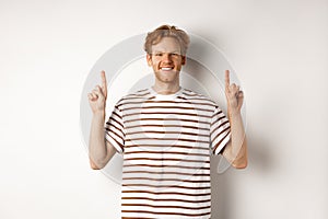 Smiling young male student with red hair showing logo, pointing fingers up and looking happy, standing over white