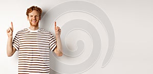 Smiling young male student with red hair showing logo, pointing fingers up and looking happy, standing over white