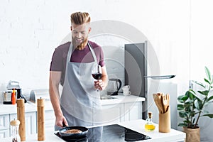 smiling young man in apron holding glass of red wine while cooking steak