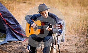 Smiling young man in black hat sitting near touristic tent and playing guitar in forest