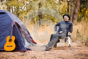 Smiling young man in black hat sitting near touristic tent and playing guitar in forest