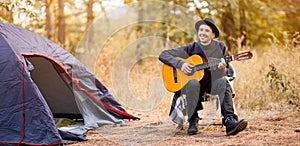 Smiling young man in black hat sitting near touristic tent and playing guitar in forest