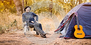 Smiling young man in black hat sitting near touristic tent and playing guitar in forest