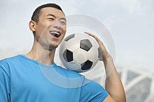 Smiling young man in a blue t-shirt holding soccer ball on his shoulder, outdoors in Beijing, China