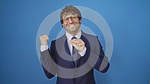 Smiling young man in business suit confidently pointing behind his back with thumbs up, presenting space on isolated blue