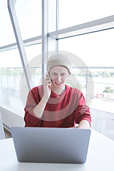 Smiling young man in the casual clothing talks on the phone and uses a laptop near the window
