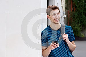 Smiling young man with cellphone and backpack leaning against wall